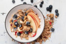 An black-rimmed white ceramic bowl filled with milk, cereal, blueberries, apple slices and almonds with an ornate silver spoon sticking out. Granola and blueberries are outside the bowl and all sitting on a painted wood surface.