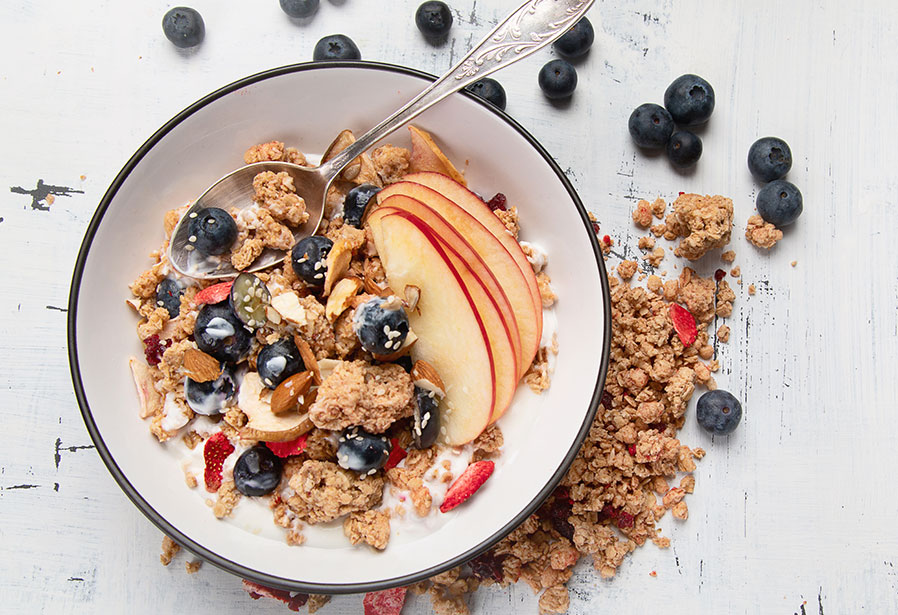 An black-rimmed white ceramic bowl filled with milk, cereal, blueberries, apple slices and almonds with an ornate silver spoon sticking out. Granola and blueberries are outside the bowl and all sitting on a painted wood surface.