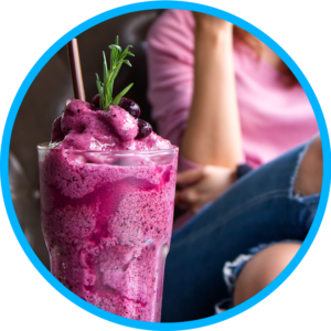 Selective focus image of a woman in ripped jeans and a dark pink top behind a berry smoothie with a straw in a glass tumbler.